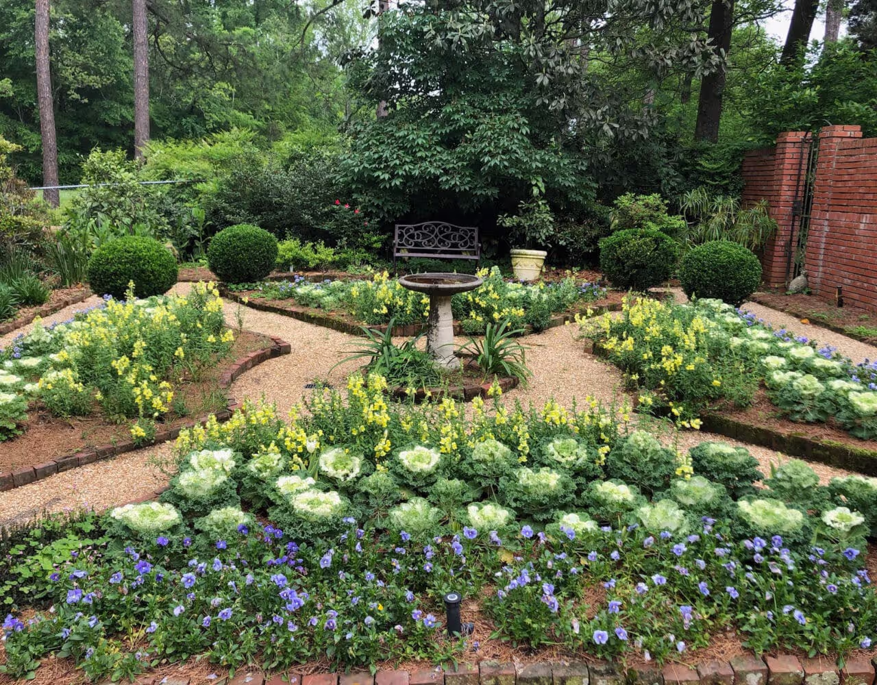 A formal garden with neatly arranged flower beds, featuring yellow and purple blooms, green foliage, a central birdbath, and a decorative bench, surrounded by a brick pathway.