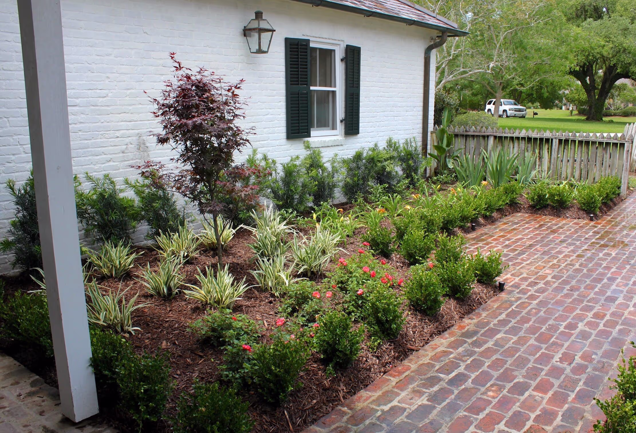 A landscaped garden beside a white brick house with green shutters, featuring a mix of shrubs, decorative grasses, and blooming flowers along a brick pathway.