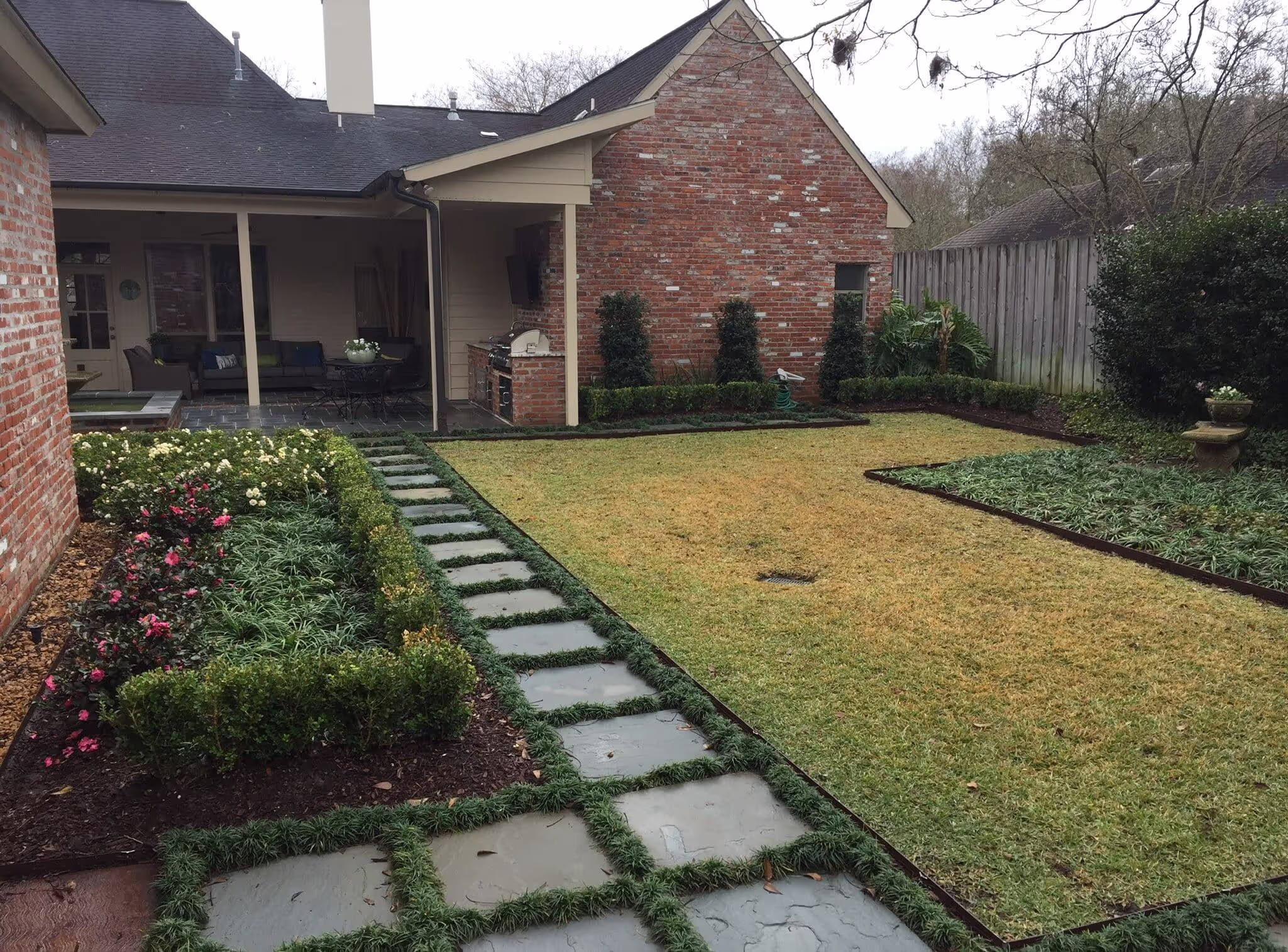 A neatly landscaped backyard with a brick house, featuring a stone pathway, trimmed hedges, flowering plants, and a well-maintained lawn.