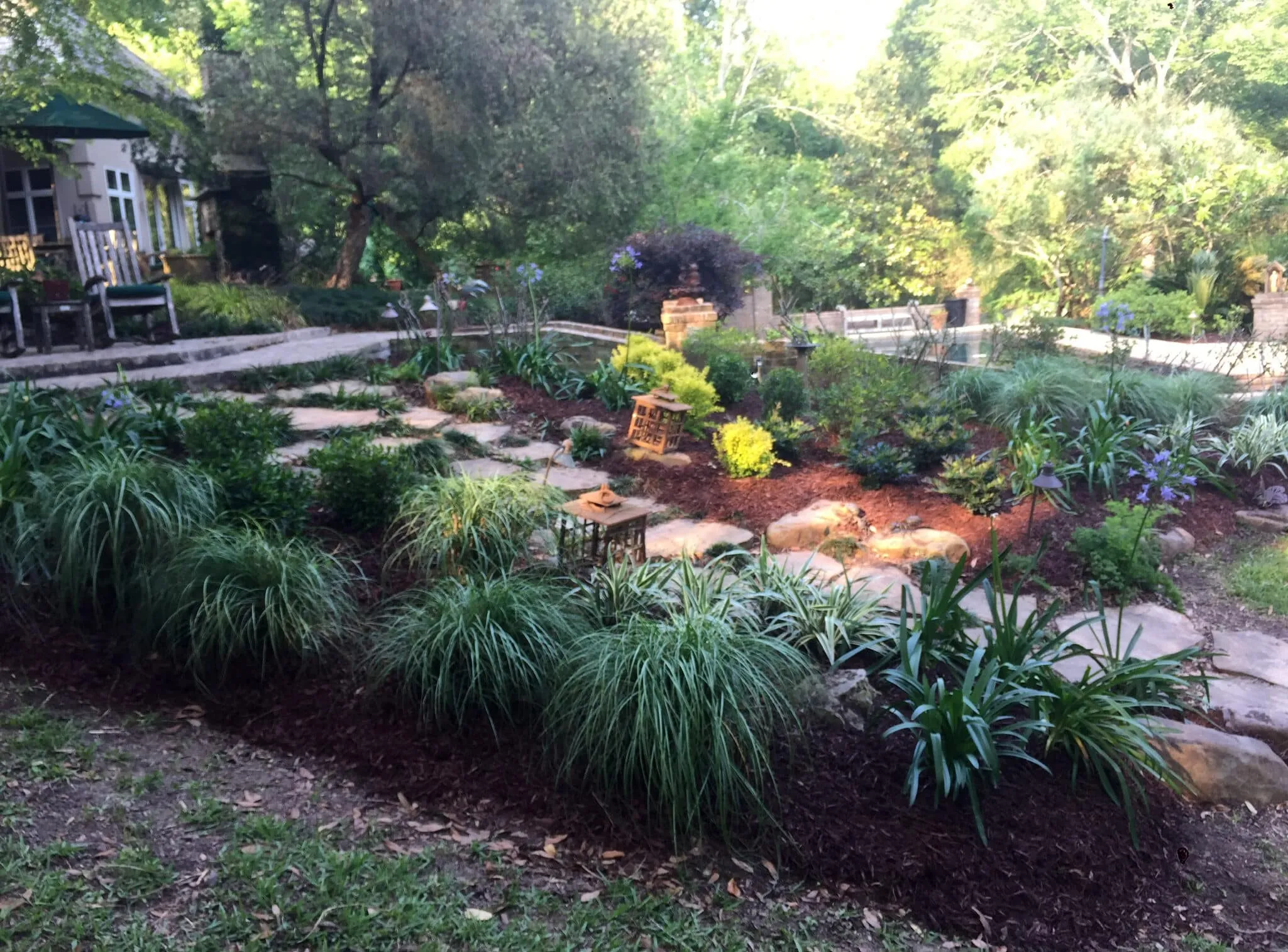 A landscaped garden with a stone pathway, lush greenery, ornamental grasses, flowering plants, and decorative lanterns, leading to a patio area with outdoor seating.