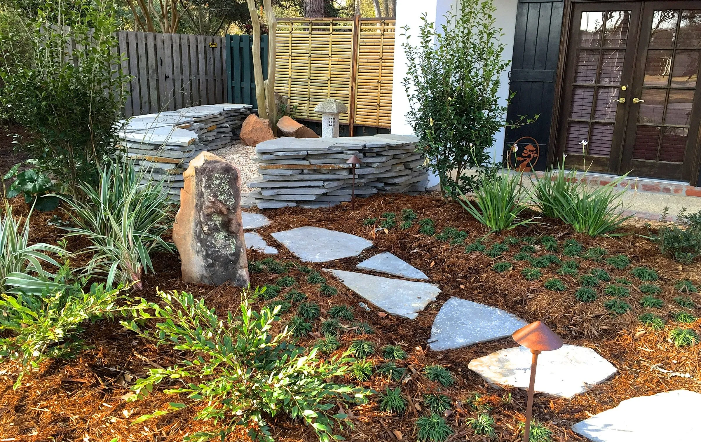A landscaped garden with a stone pathway, various shrubs, ornamental grasses, and a rock feature, leading to a patio area with stacked stone walls and a bamboo privacy screen.