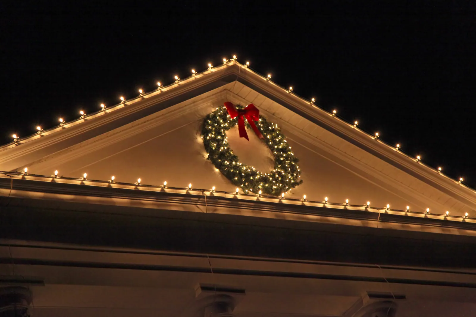 Roofline lights and a lit wreath with a red bow on a house gable at night in Walker, LA